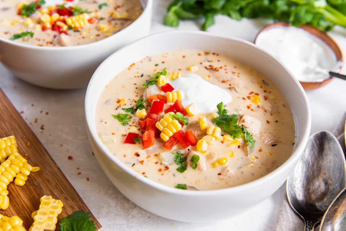Two bowls of chicken corn chowder topped with sour cream and bell peppers next to a cutting board with corn kernels on it.