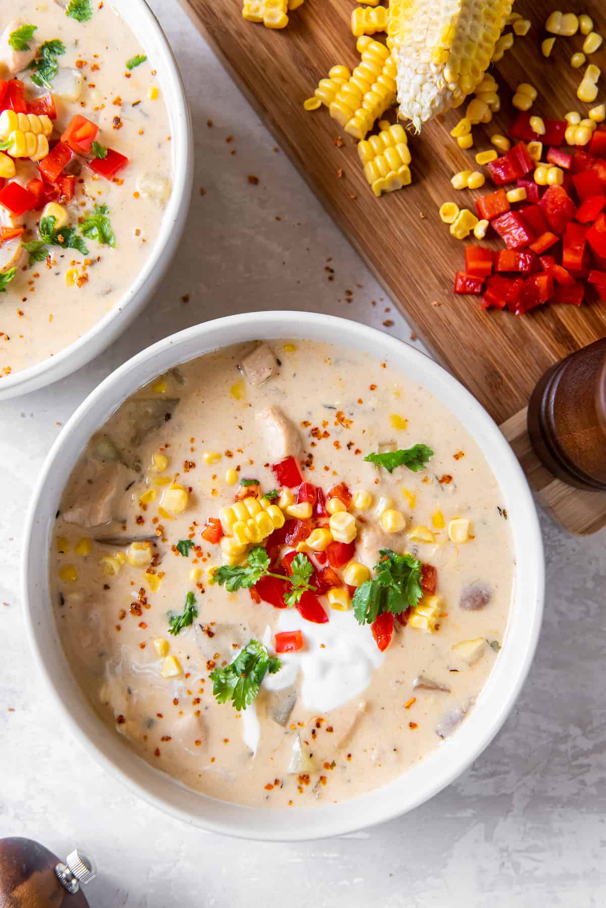 A top down shot of two white bowls filled with chowder with chicken and corn topped with sour cream and diced bell peppers.