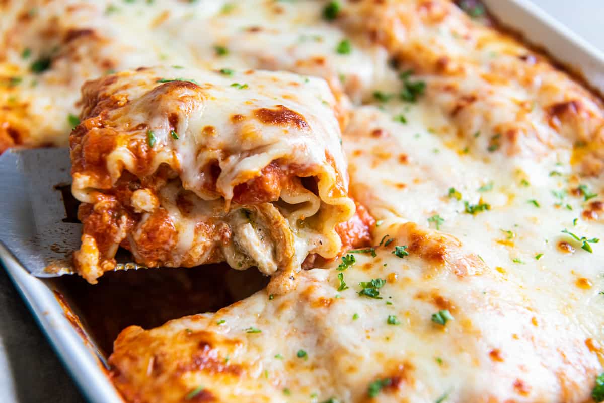 A spatula lifting a cheesy chicken lasagna rollup from a baking dish.