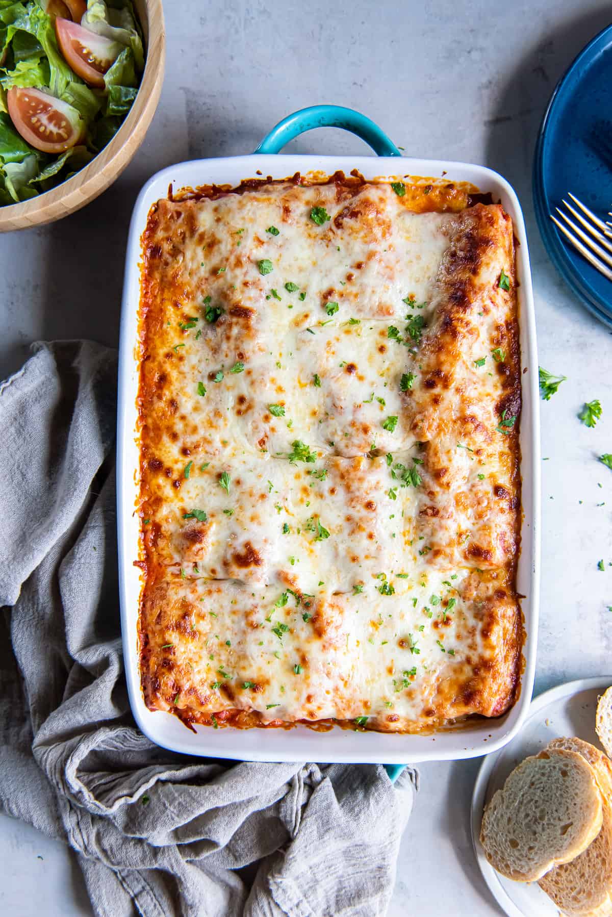 A top down shot of lasagna rollups topped with sauce and melted cheese i a baking dish next to a green salad and slices of bread.