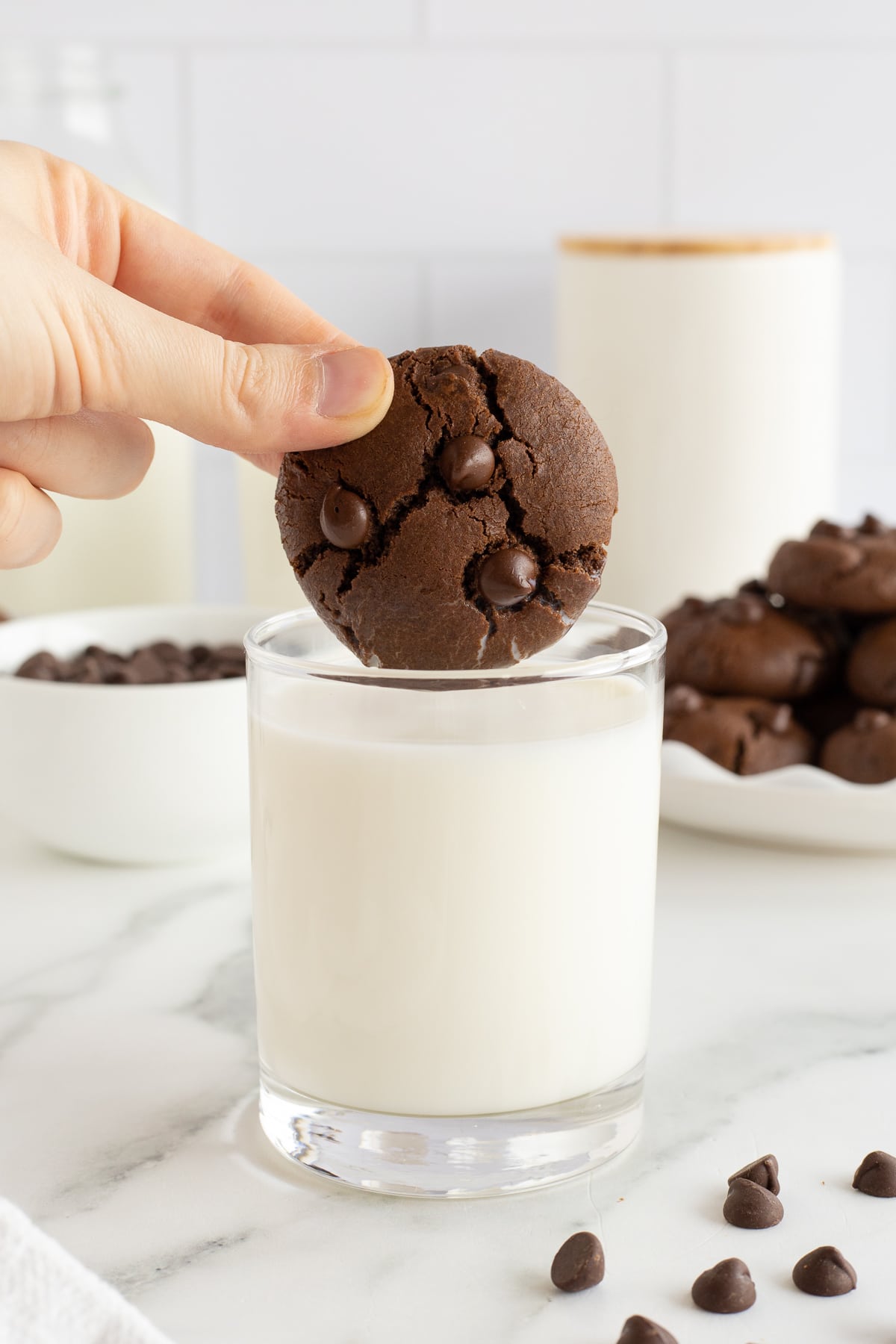 A hand dipping a chocolate cookie with chocolate chips into a glass of milk.