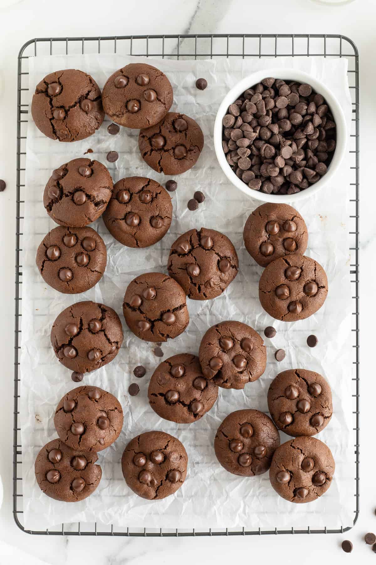 A top down shot of double chocolate cookies on a wire rack with a small bowl of chocolate chips.