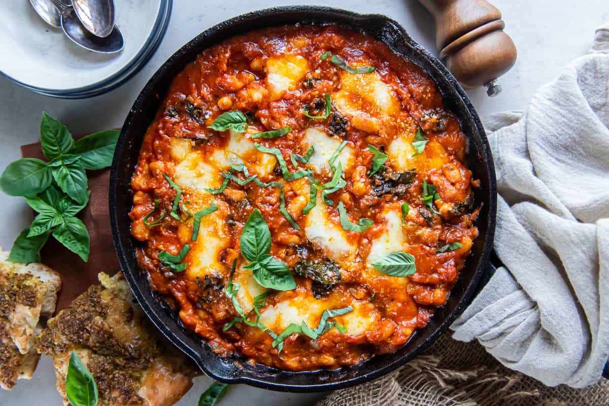 Cannellinni beans in marinara sauce with kale and cheese in a cast iron skillet next to pieces of foccacia bread and fresh basil.