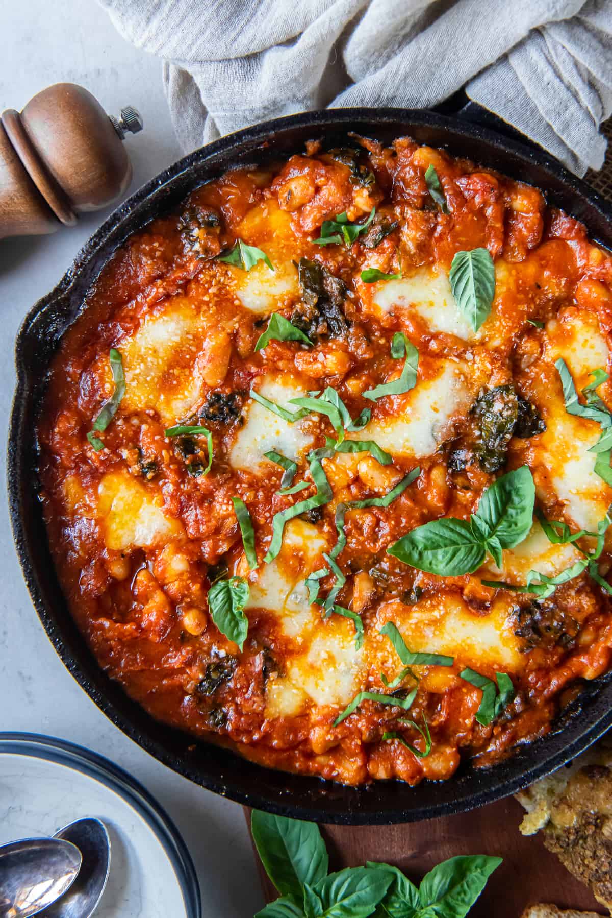 A top down shot of a cast iron skillet filled with white beans in marinara sauce with kale and mozzarella.