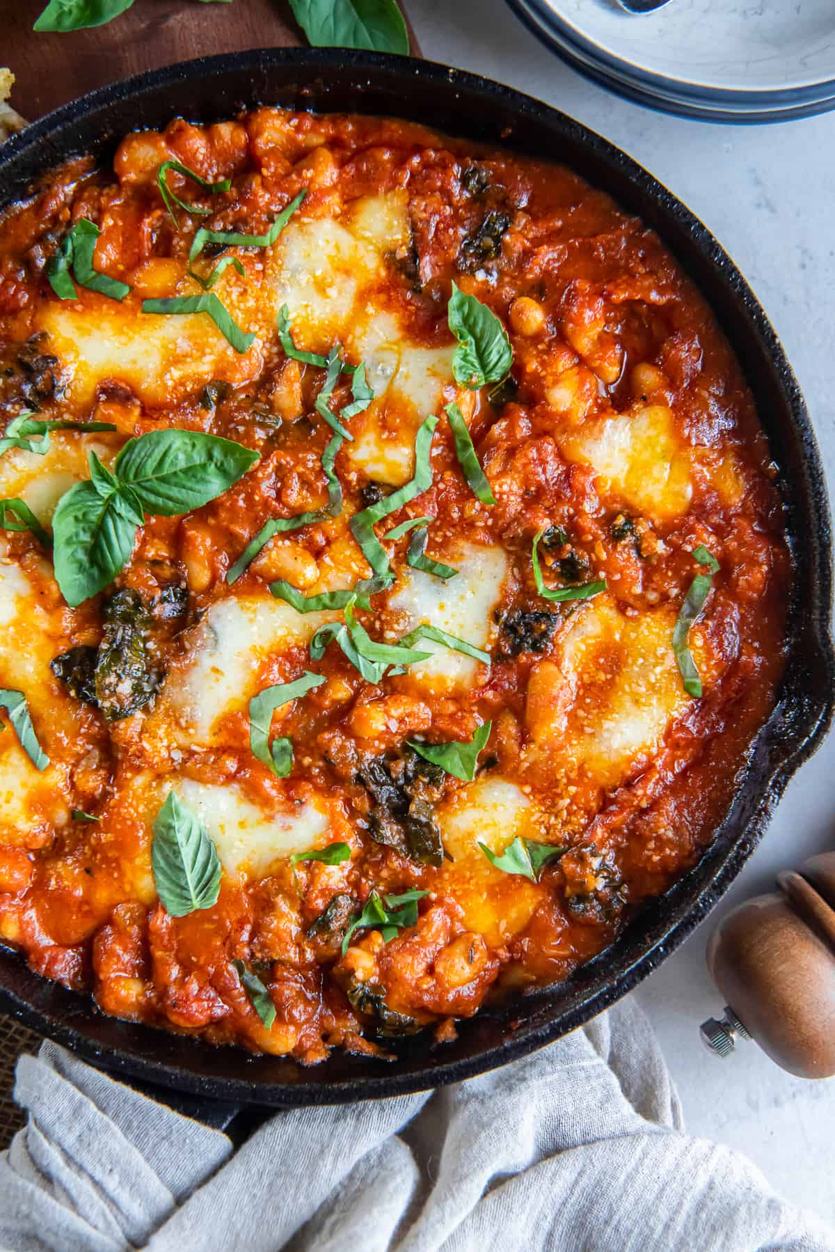 A top down shot of a cast iron skillet filled with white beans in marinara sauce with kale and mozzarella.