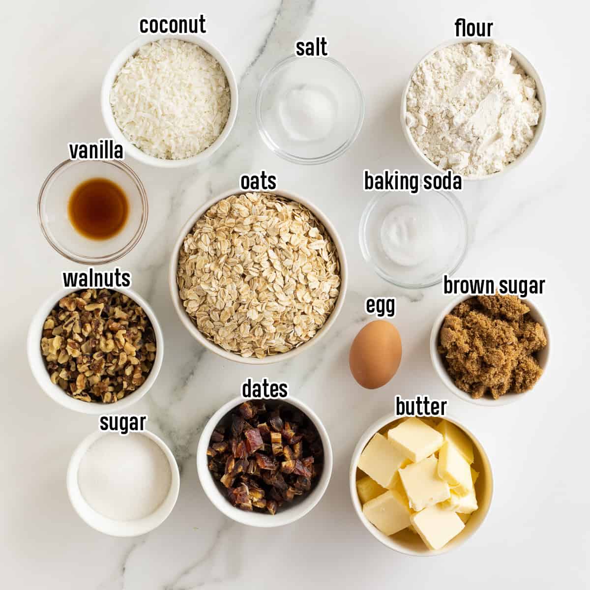 A top down shot of oats, flour, sugar, dates, and other ingredients in bowls with text.