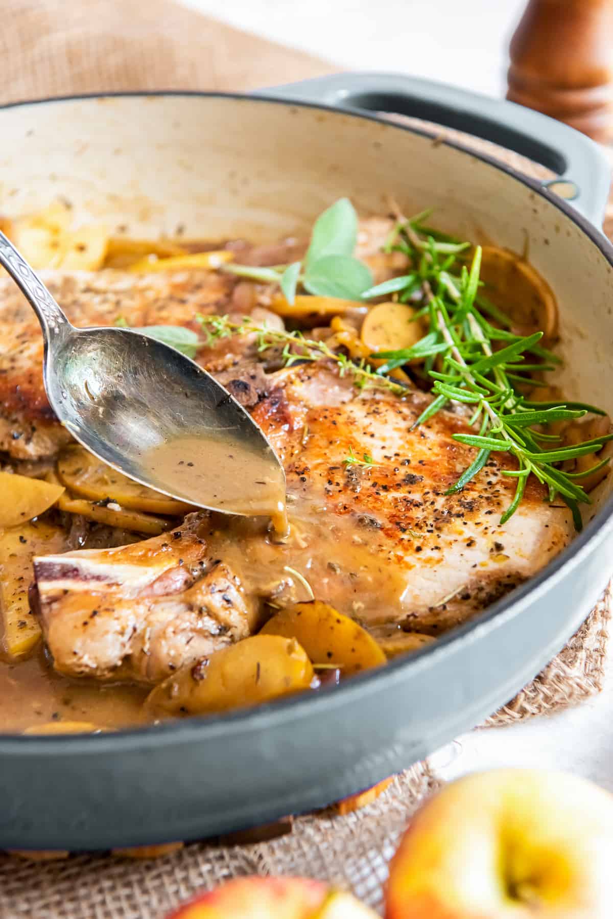 A spoon pouring pan sauce of pork chops in a skillet with apples and onions.