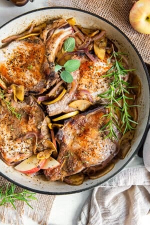 A top down shot of four pork chops in a skillet with apples and onions topped with fresh sage and sprigs of rosemary.