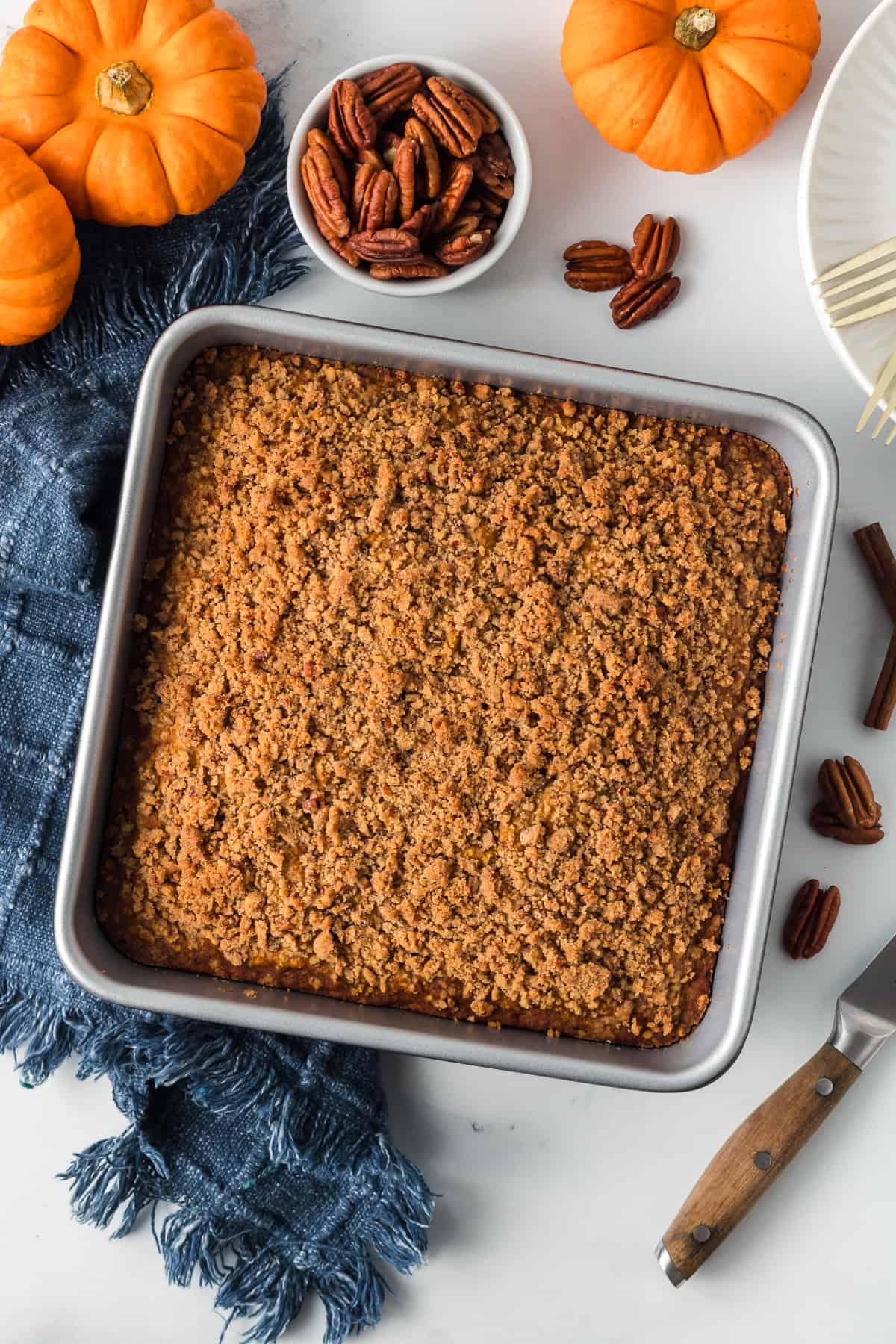 A top down shot of pumpkin crumb cake in a metal baking pan surrounded by mini pumpkins and pecans.