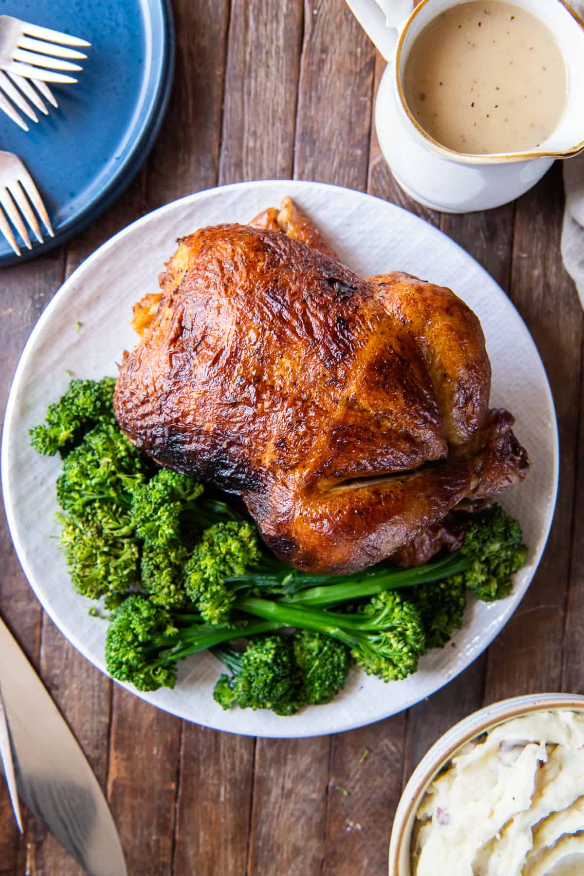 A top down shot of a rotisserie chicken on a white platter with fresh parsley and a gravy boat behind it.