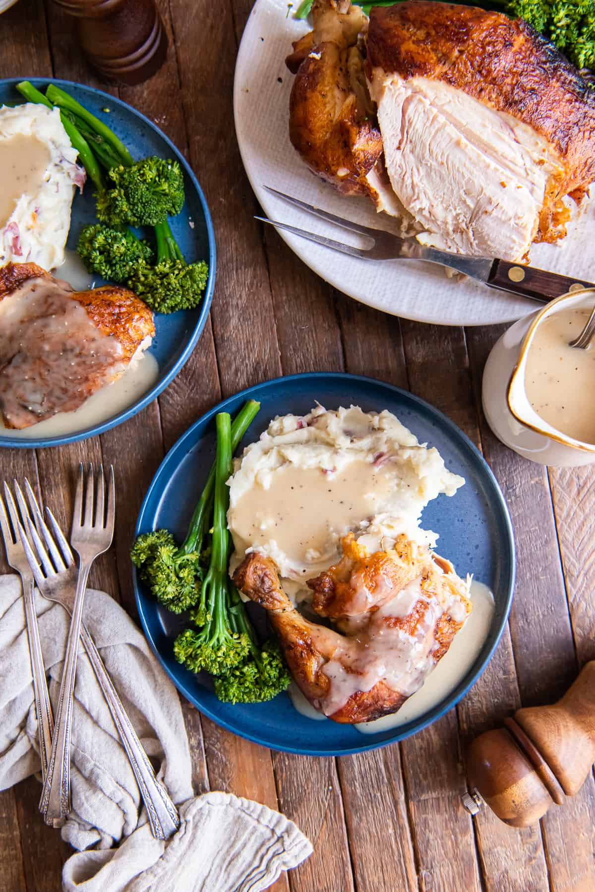 A top down shot of a roast chicken dinner on a blue plate next to a platter with a whole sliced chicken and gravy.