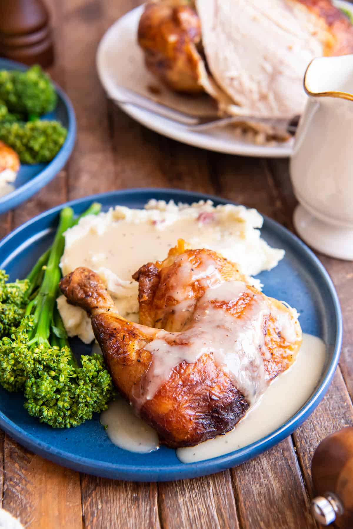Pieces of rotisserie chicken topped with gravy on a blue plate with mashed potatoes and broccolini.