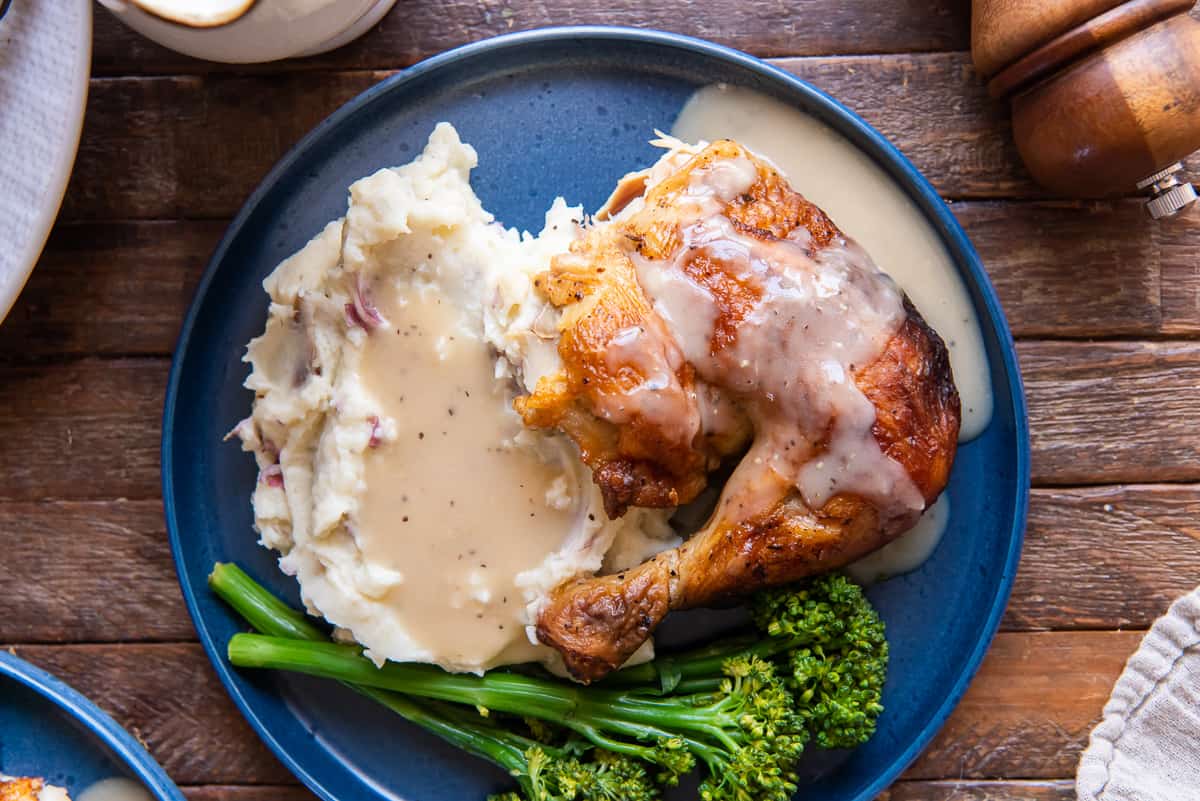 A top down shot of chicken and mashed potatoes with gavy on a blue plate with broccolini.