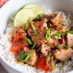 A serving of Asian pork with peppers over rice with lime wedges in a small white bowl.