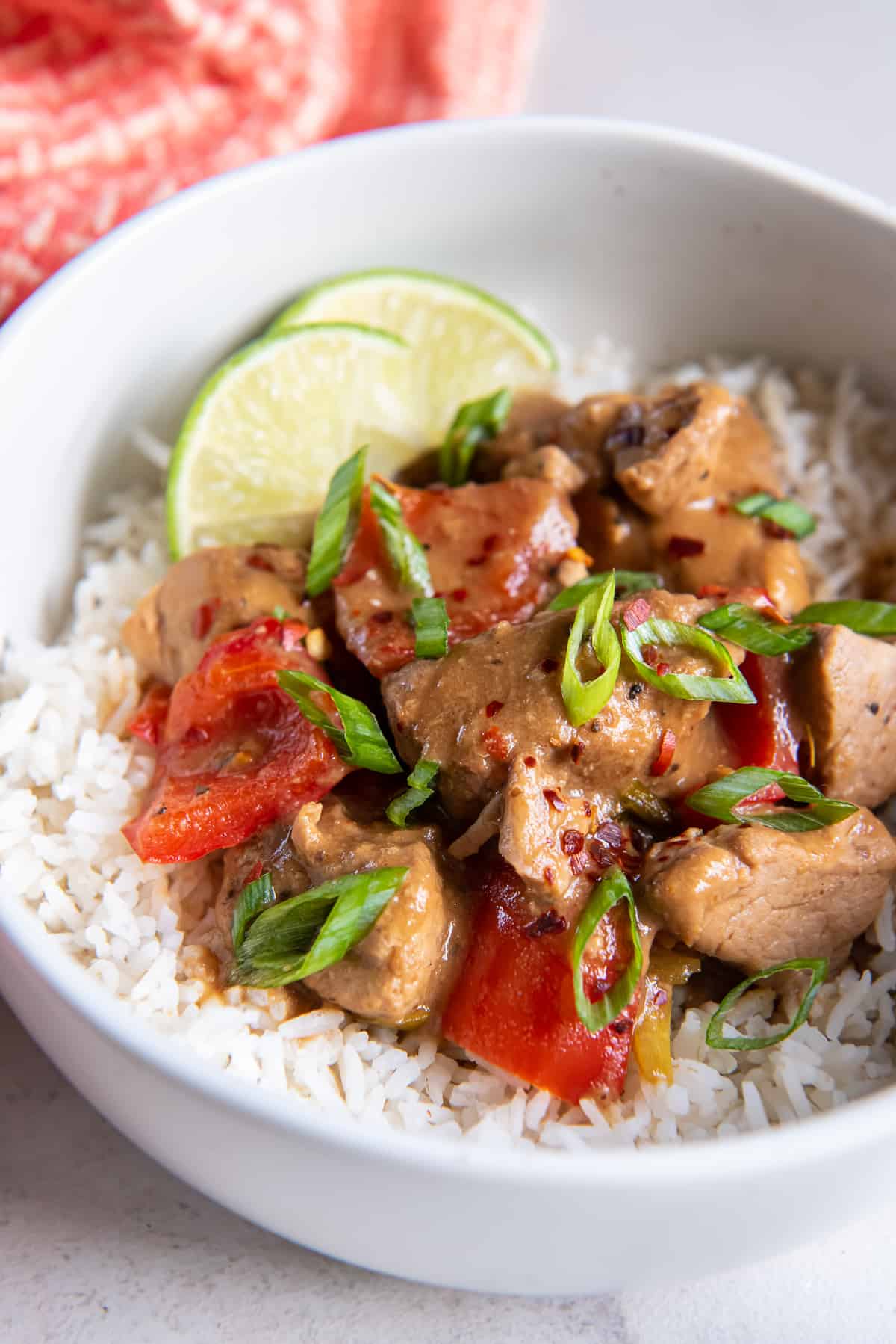 A serving of Asian pork with peppers over rice with lime wedges in a small white bowl.