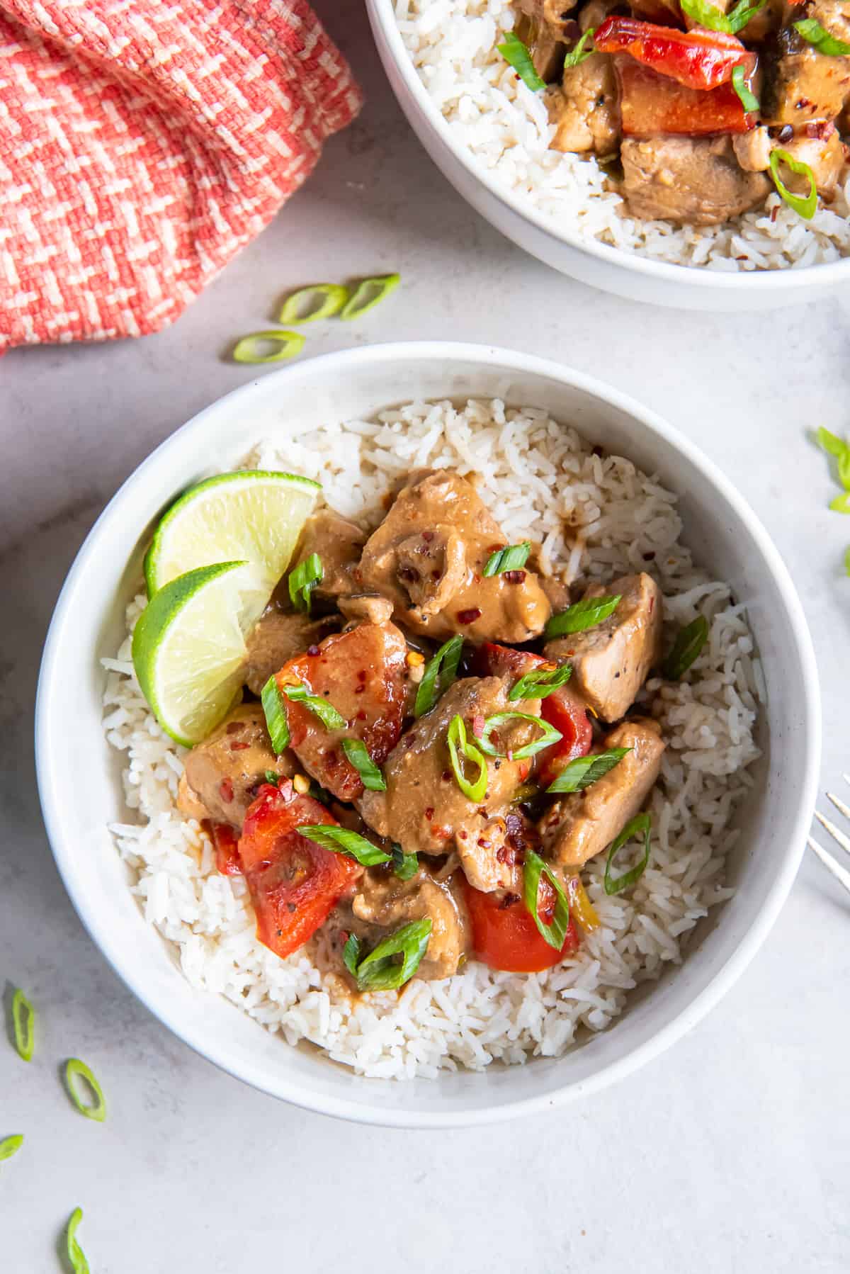 A top down shot of Asian pork with peppers over rice with lime wedges in a small white bowl next to an orange and white checked cloth.