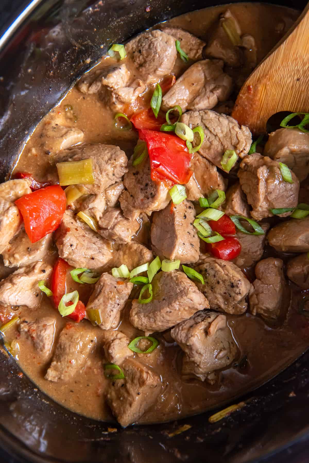 A top down shot of chunks of pork with red bell peppers and green onions in an Asian sauce in a slow cooker.