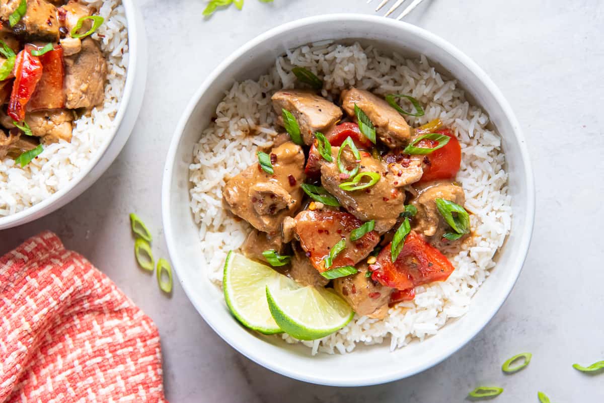 A top down shot of Asian pork with peppers over rice with lime wedges in a small white bowl next to an orange and white checked cloth.