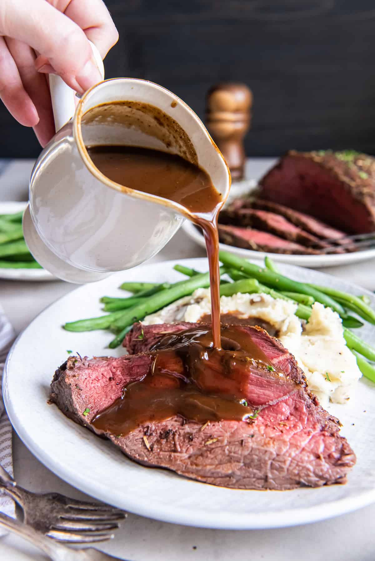 A hand pouring gravy from a gravy boat over slices of cross rib roast on a dinner plate.