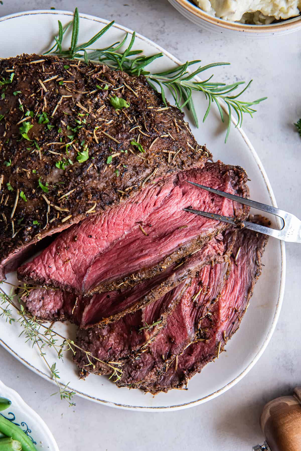 A top down shot of a sliced cross rib roast on a white platter with sprigs of fresh rosemary.