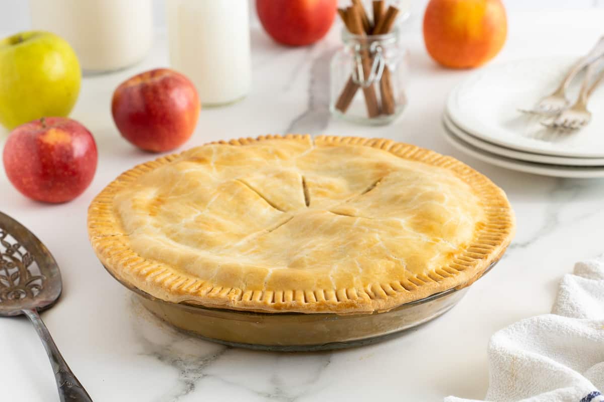 A double crust pie on a kitchen counter with apples behind it.