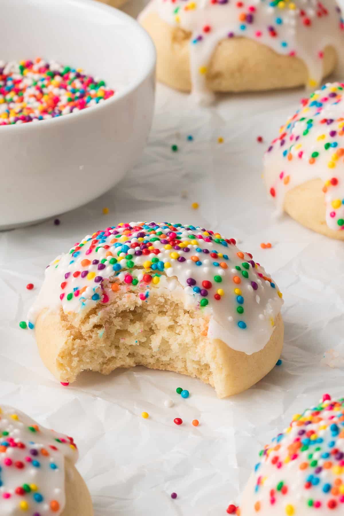 An Italian Cookie with icing and rainbow sprinkles with a bite missing on parchment papper.