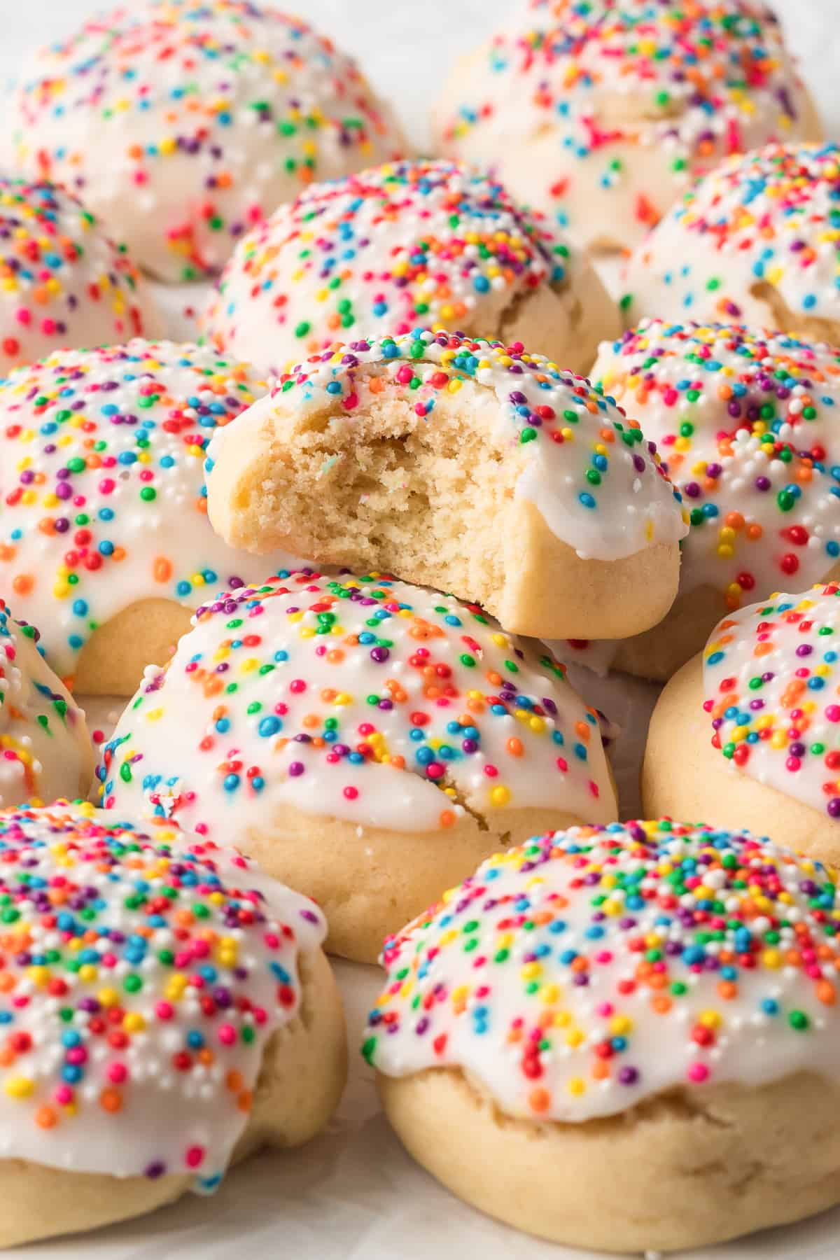 A close up of a pile of Italian Cookies with icing and rainbow sprinkles with a bite missing from the cookie on top.