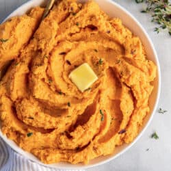 A top down shot of a bowl of mashed sweet potatoes topped with thyme and butter next to a pepper shaker an sprigs of fresh thyme.