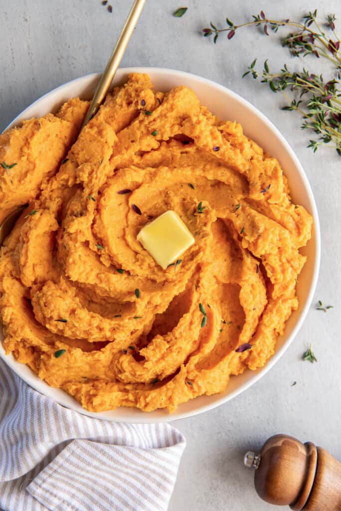 A top down shot of a bowl of mashed sweet potatoes topped with thyme and butter next to a pepper shaker an sprigs of fresh thyme.