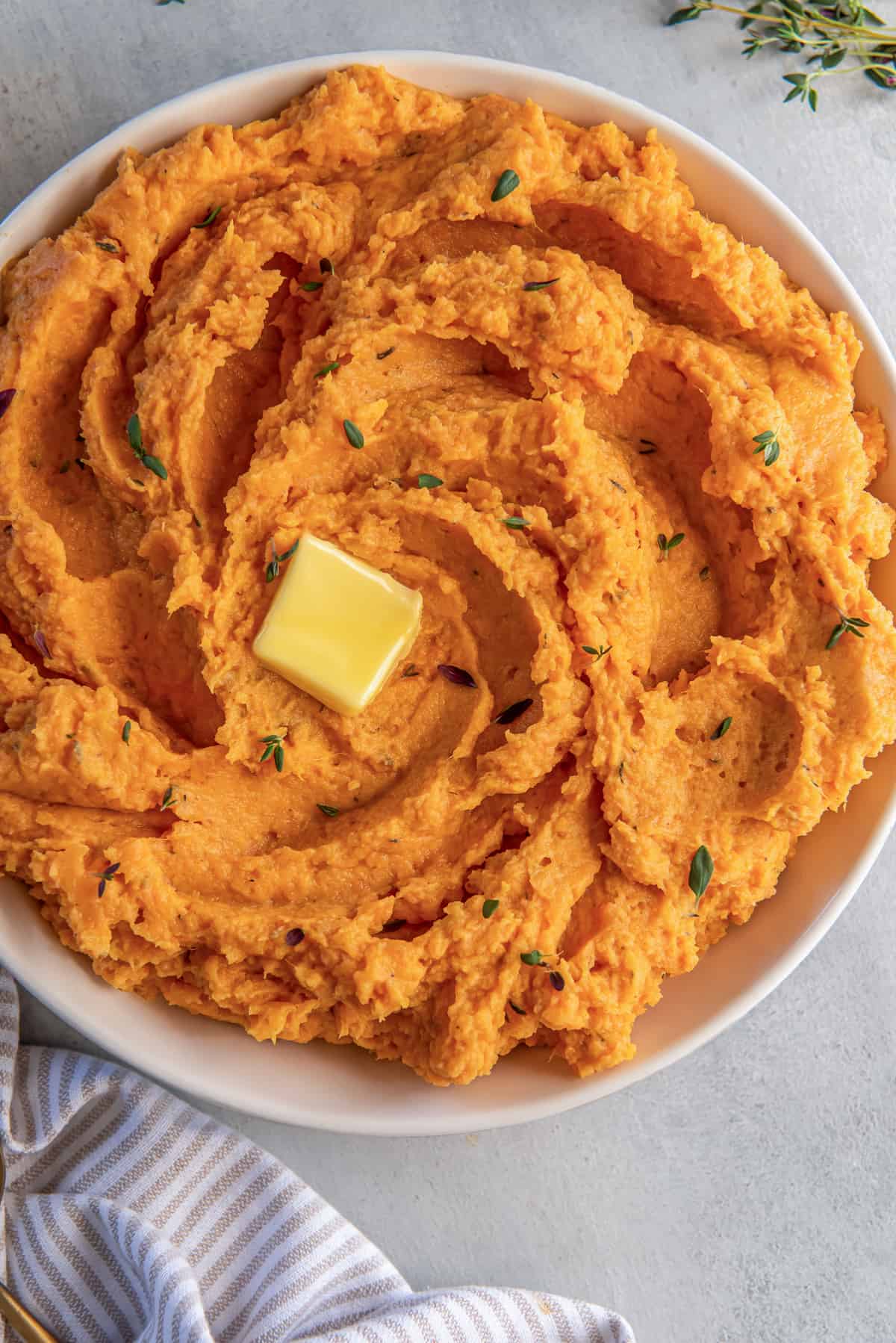 A top down shot of a bowl of mashed sweet potatoes topped with thyme and butter.