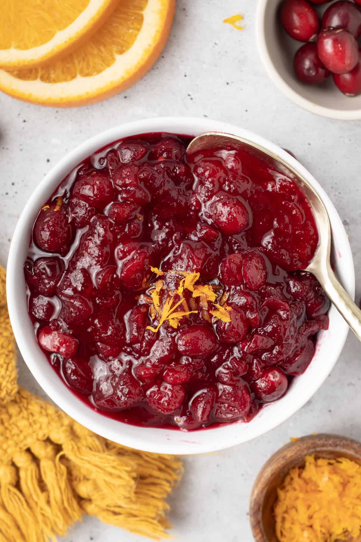 A top down shot of a spoon resting in a bowl of cranberry sauce next to a gold cloth and orange slices.