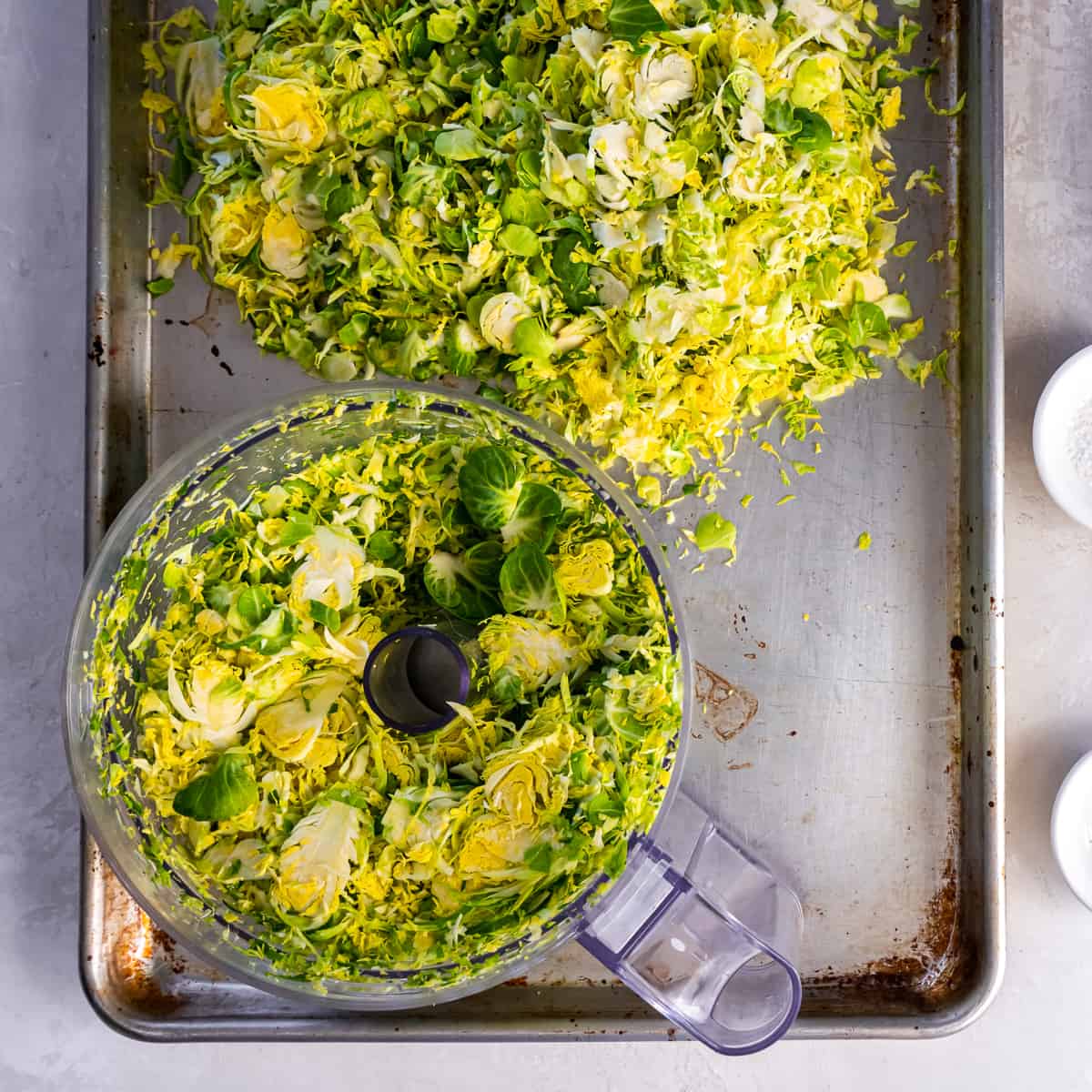 Shredded Brussels sprouts in the bowl of a food processor and scattered over a baking sheet.