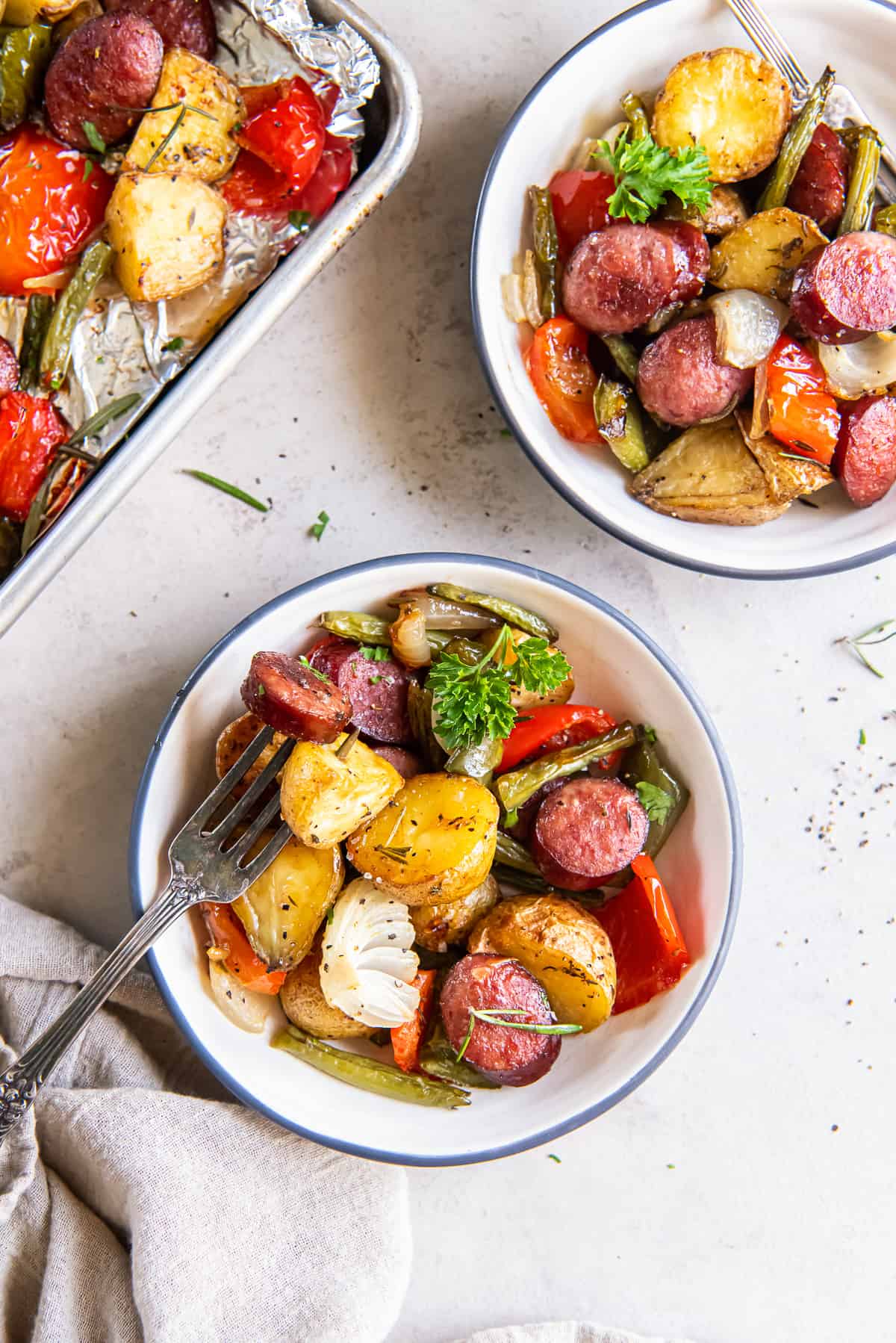 Two bowls filled with roasted sausage and potatoes with green beans and bell peppers.