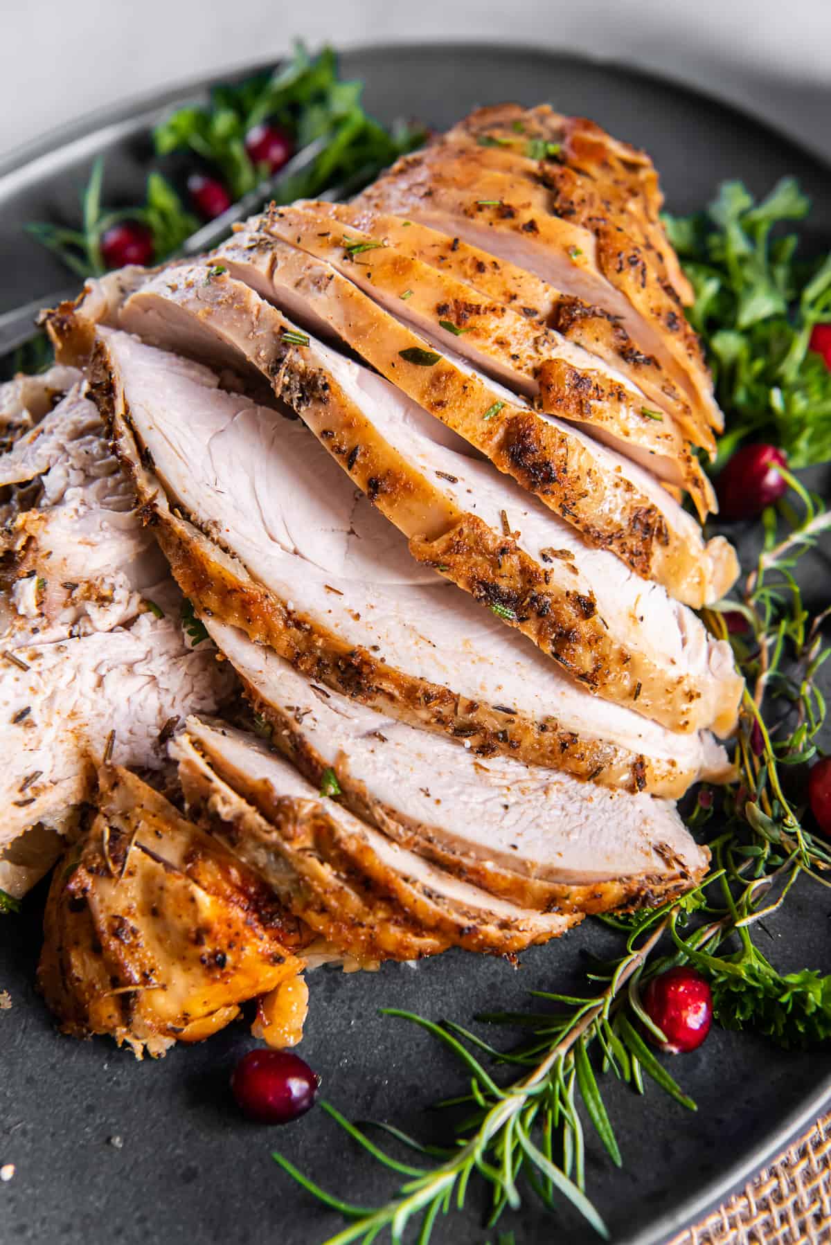 A top down shot of a sliced turkey breast on a grey platter with cranberries and fresh herbs.