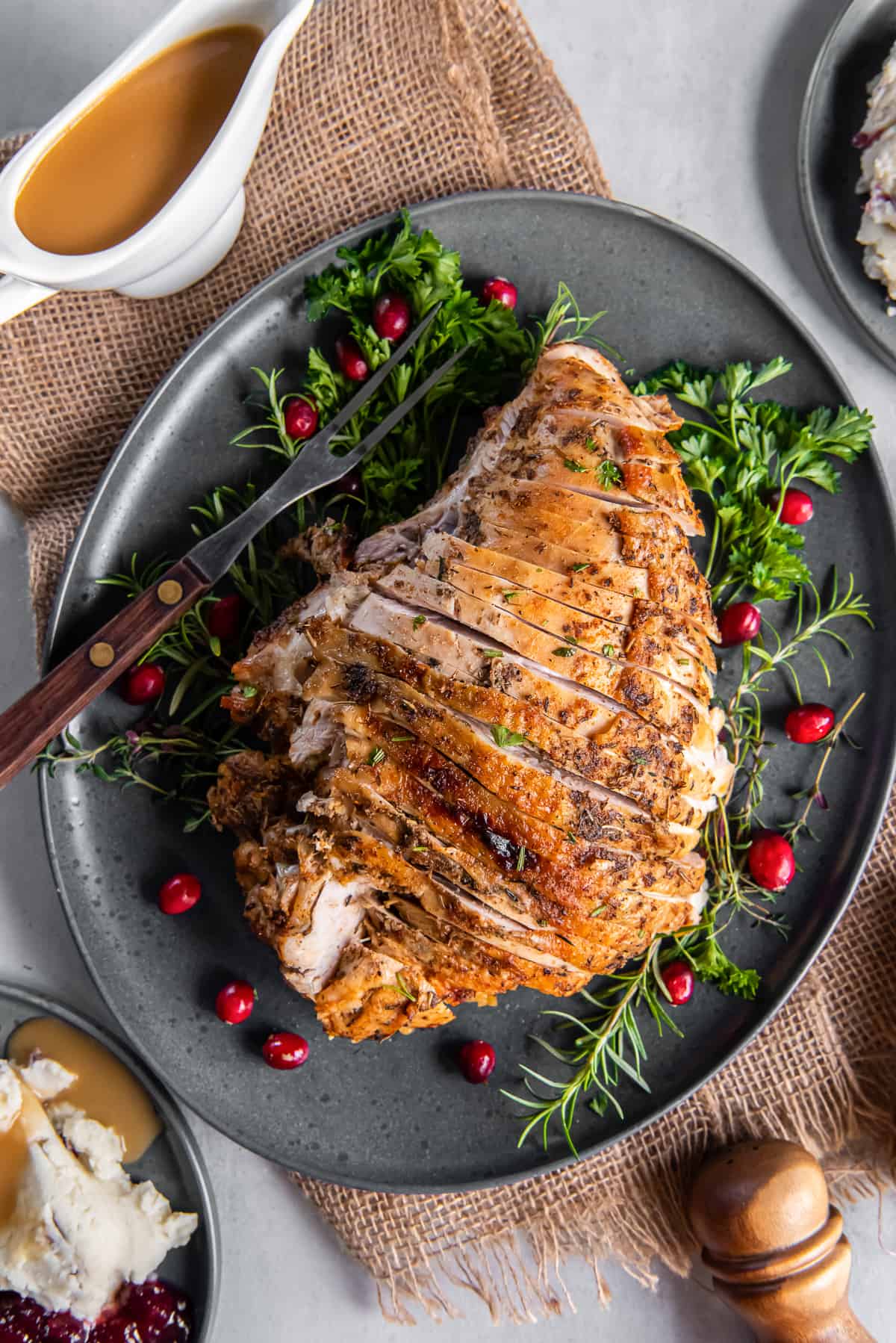 A top down shot of a sliced turkey breast on a grey platter with fresh herbs and cranberries next to a gravy boat.