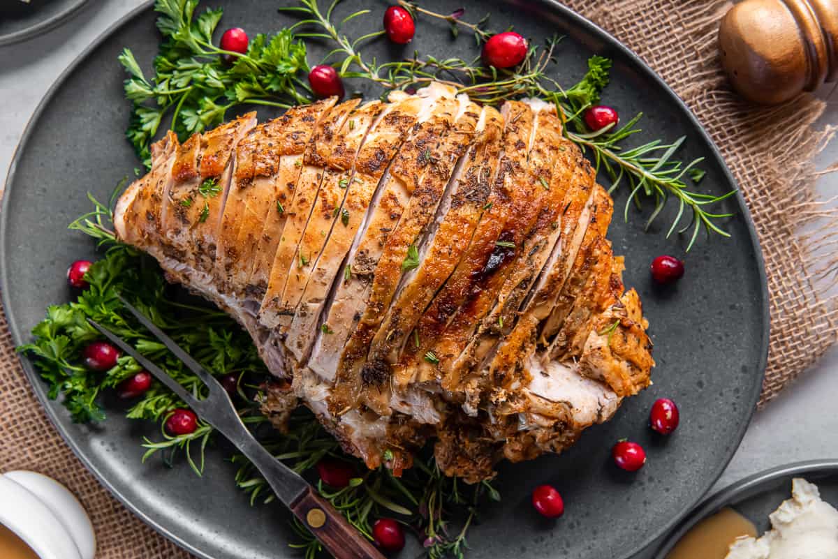 A top down shot of a sliced slow cooker turkey breast on a grey platter with cranberries and fresh herbs.