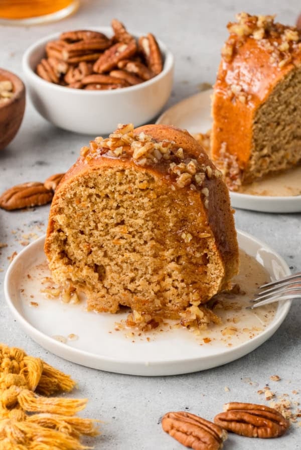 Two slices of sweet potato bundt cake on white plates next to a small bowl of pecans.