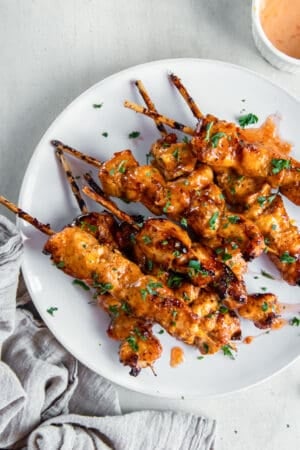 A top down shot of bang bang chicken skewers on a white plate next to a grey cloth,