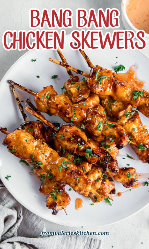 A top down shot of bang bang chicken skewers on a white plate next to a grey cloth with text.