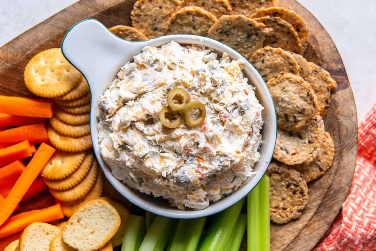 A top down shot of a bowl of olive dip on a wood platter with veggies and crackers.