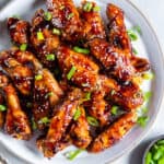 Glazed Asian chicken wings on a platter next to a small bowl of sliced green onions.