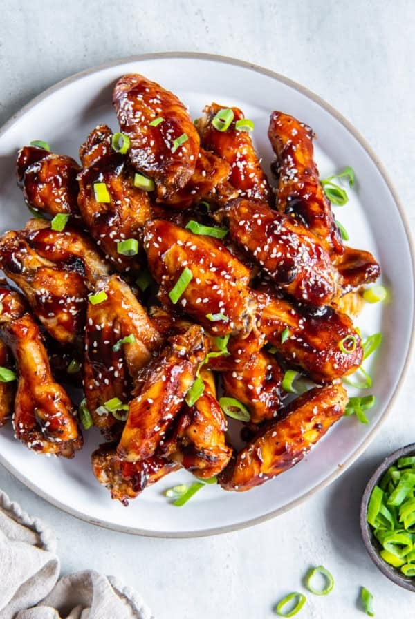Glazed Asian chicken wings on a platter next to a small bowl of sliced green onions.