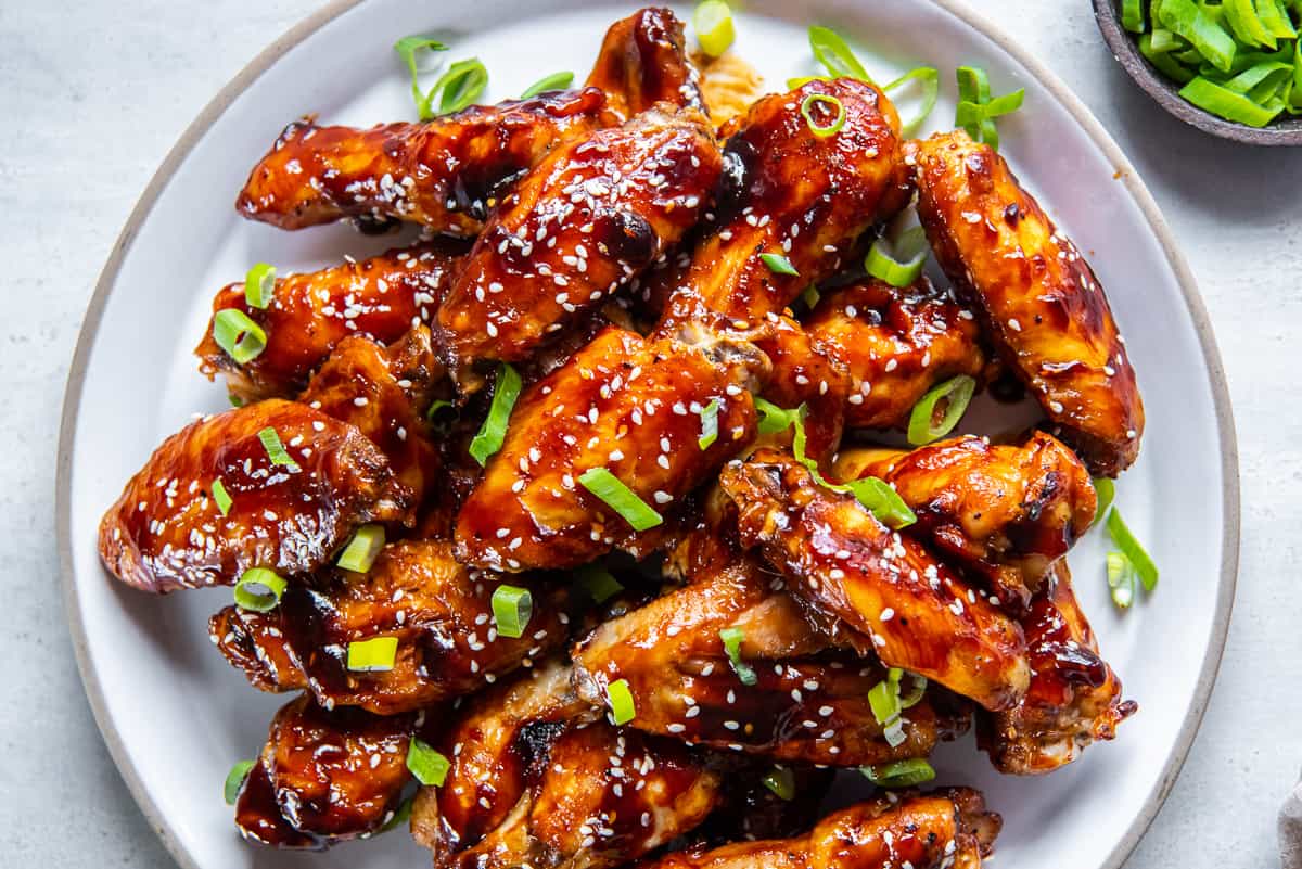 A pile of sticky asian wings on a white platter topped with sesame seeds and sliced green onions.