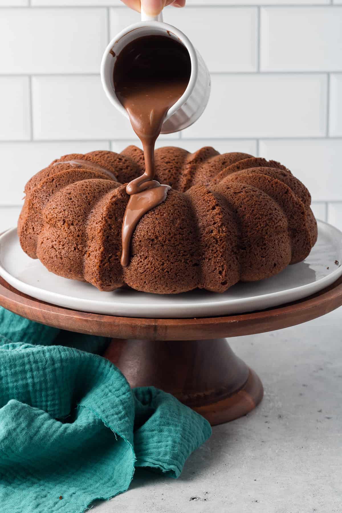 Chocolate kahlua glaze pouring from a white cup over a bundt cake on a cake pedestal.