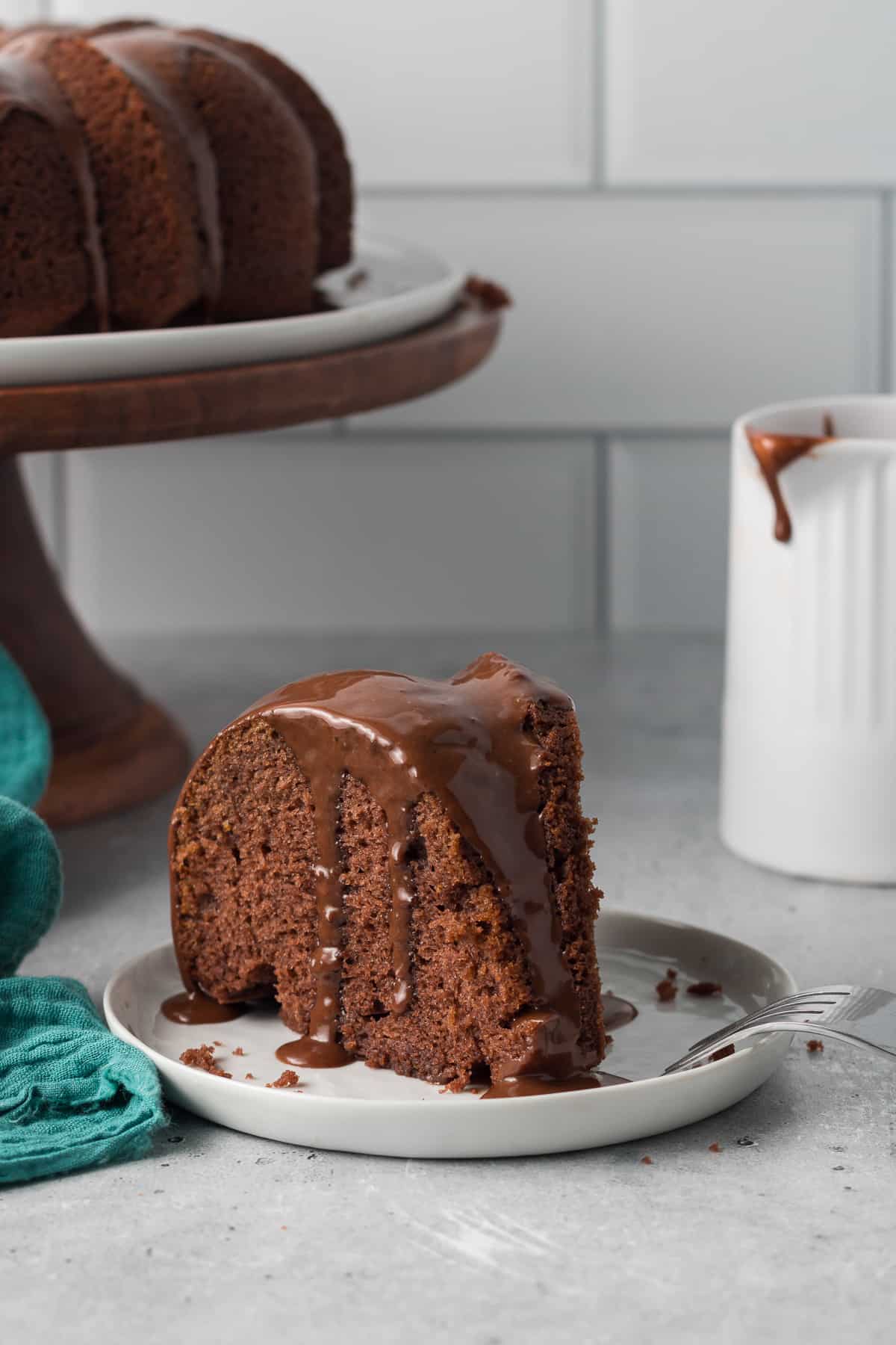 A slice of black russian cake on a white plate in front of a cake pedestal holding a bundt cake.