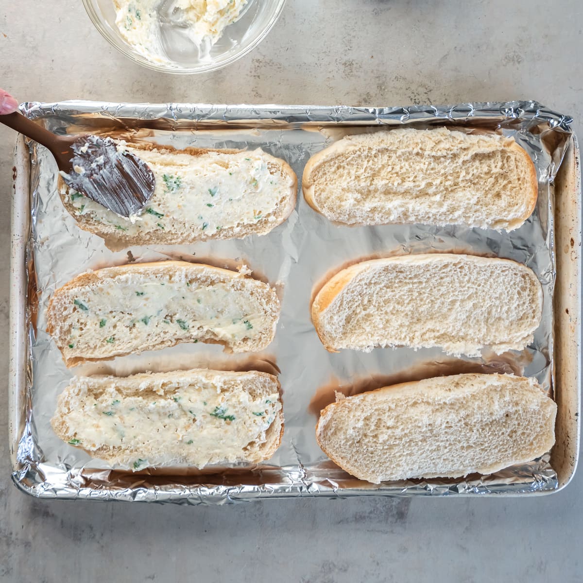 A pastry brush spreading garlic butter on a hoagie roll on a baking sheet.