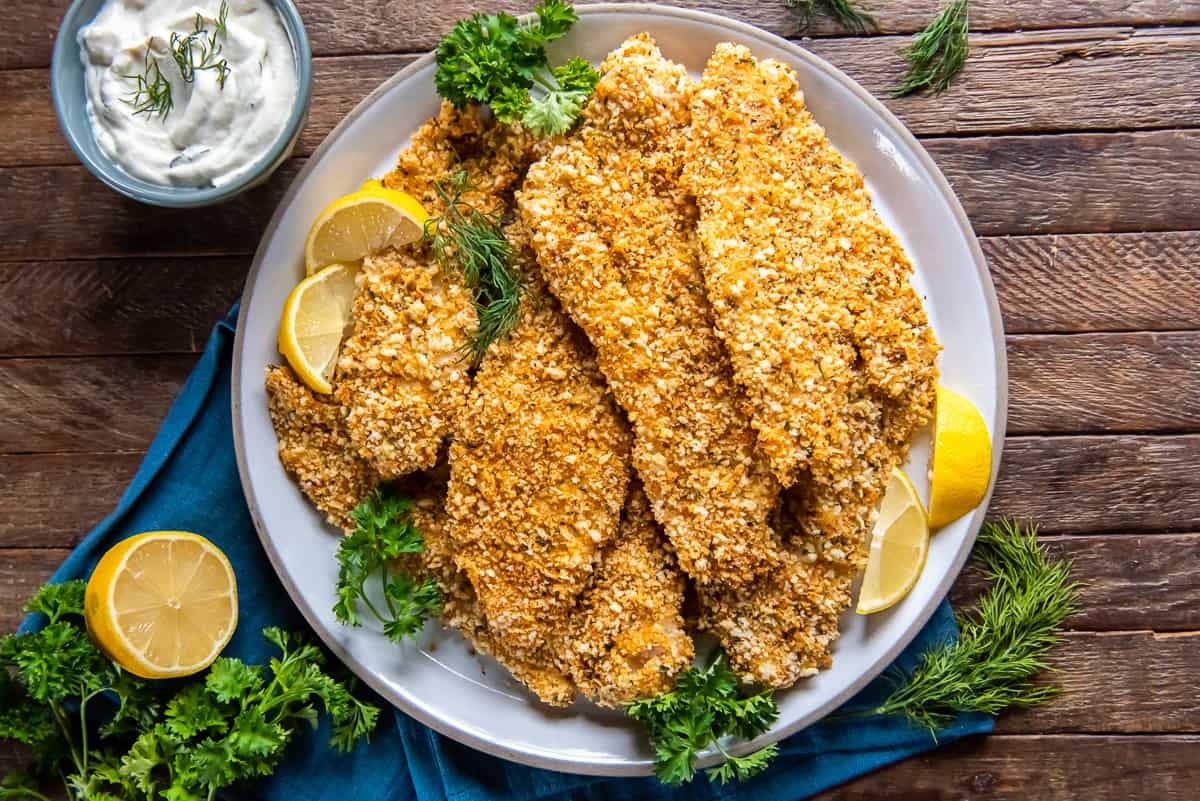 A top down shot of a white plate full of crispy baked fish with lemon wedges and parsley.