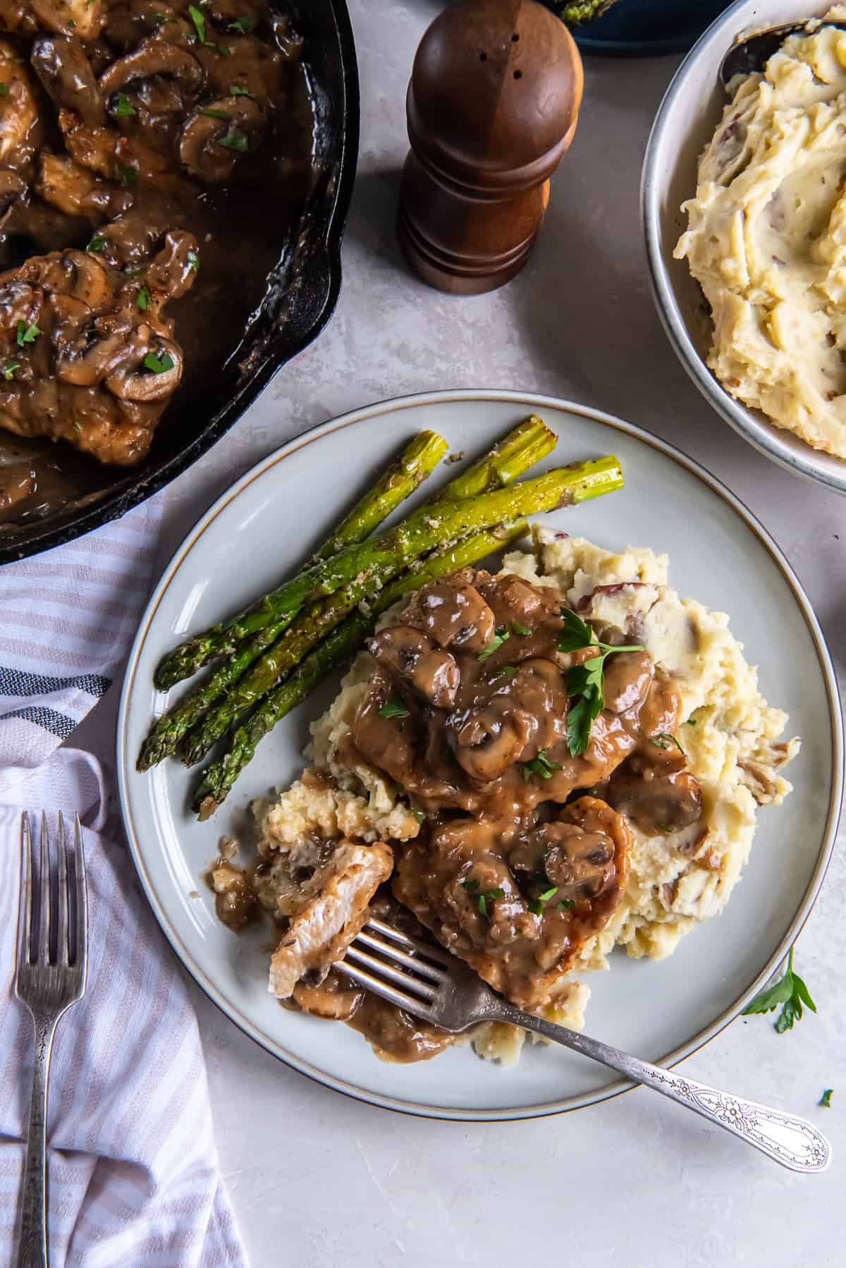 A top down shot of a fork resting on a plate of chicken marsala with asparagus and mashed potatoes.