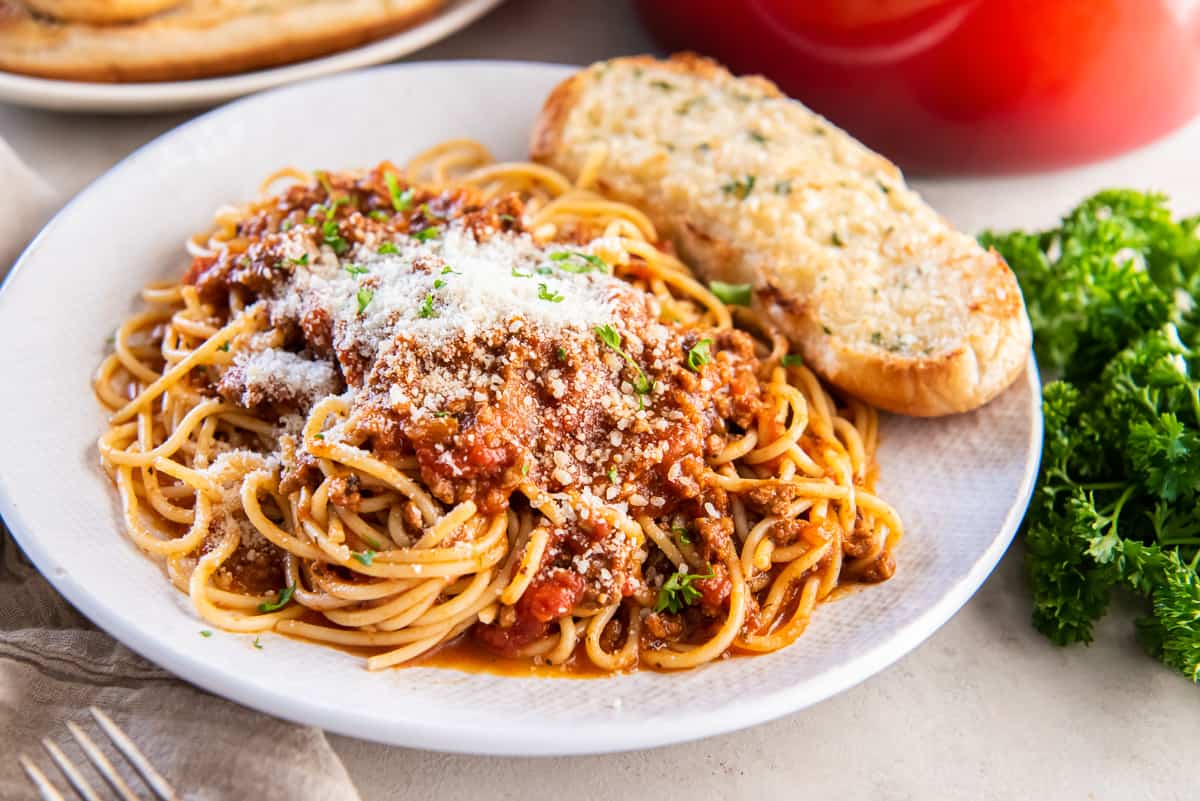 Spaghetti with sauce topped with Parmesan cheese on a white plate with garlic bread.