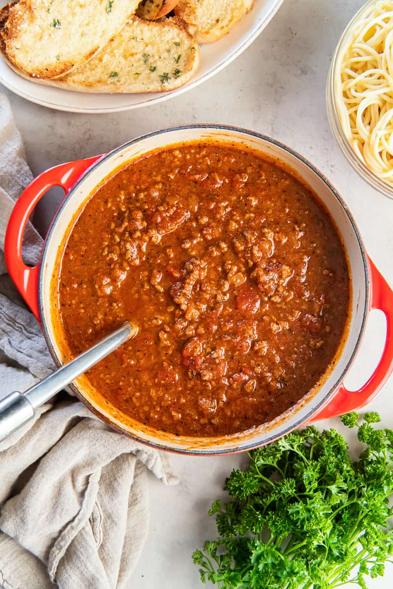 A top down shot of spaghetti sauce in a Dutch oven next to garlic bread and cooked pasta.